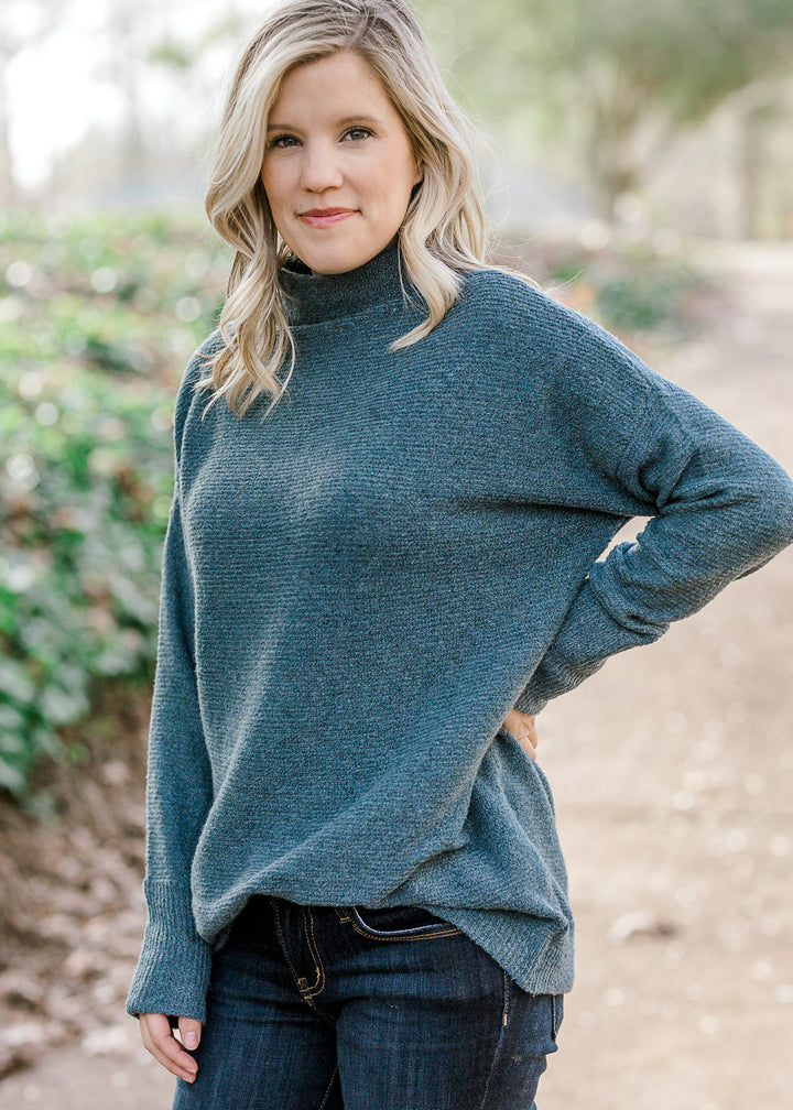 Blonde model wearing a teal sweater with a mocked neck. 
