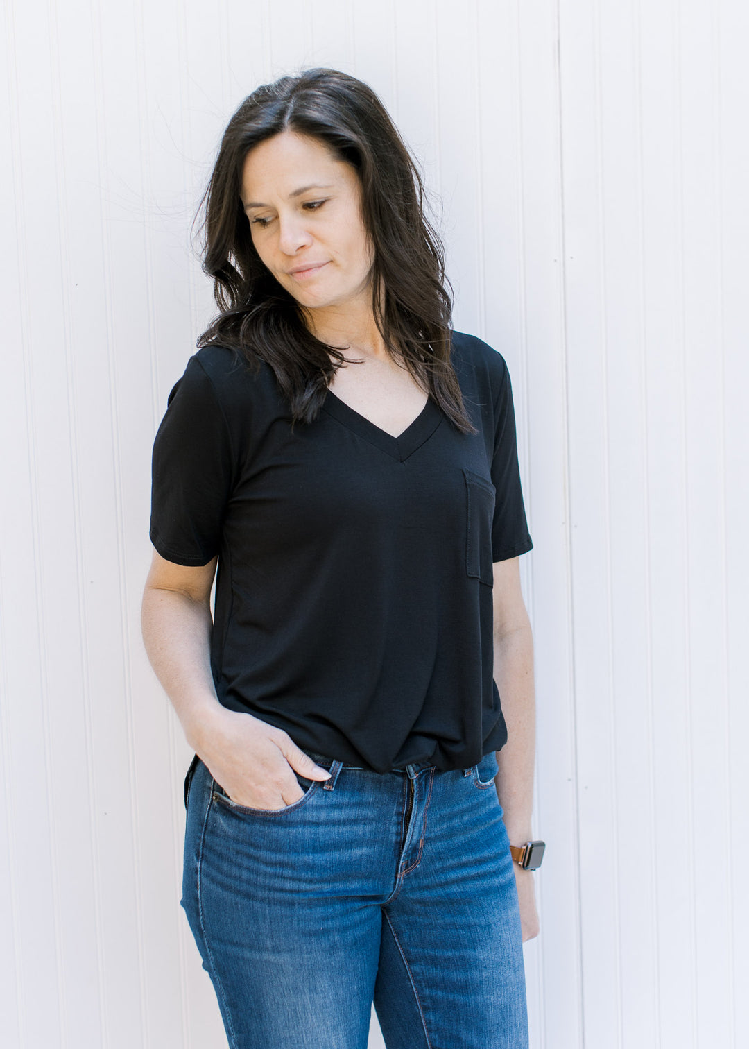 Model wearing jeans with a black, v-neck, short sleeve top with a chest pocket. 