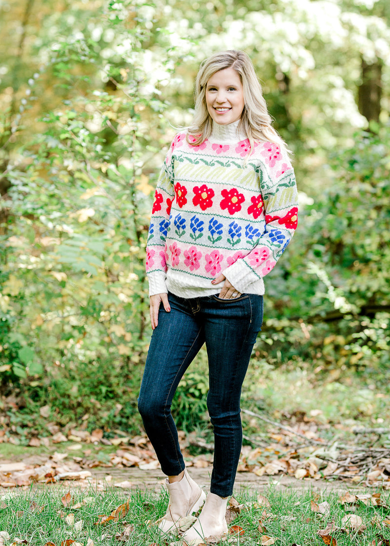 Blonde model wearing cream sweater with bright floral pattern with jeans and booties.