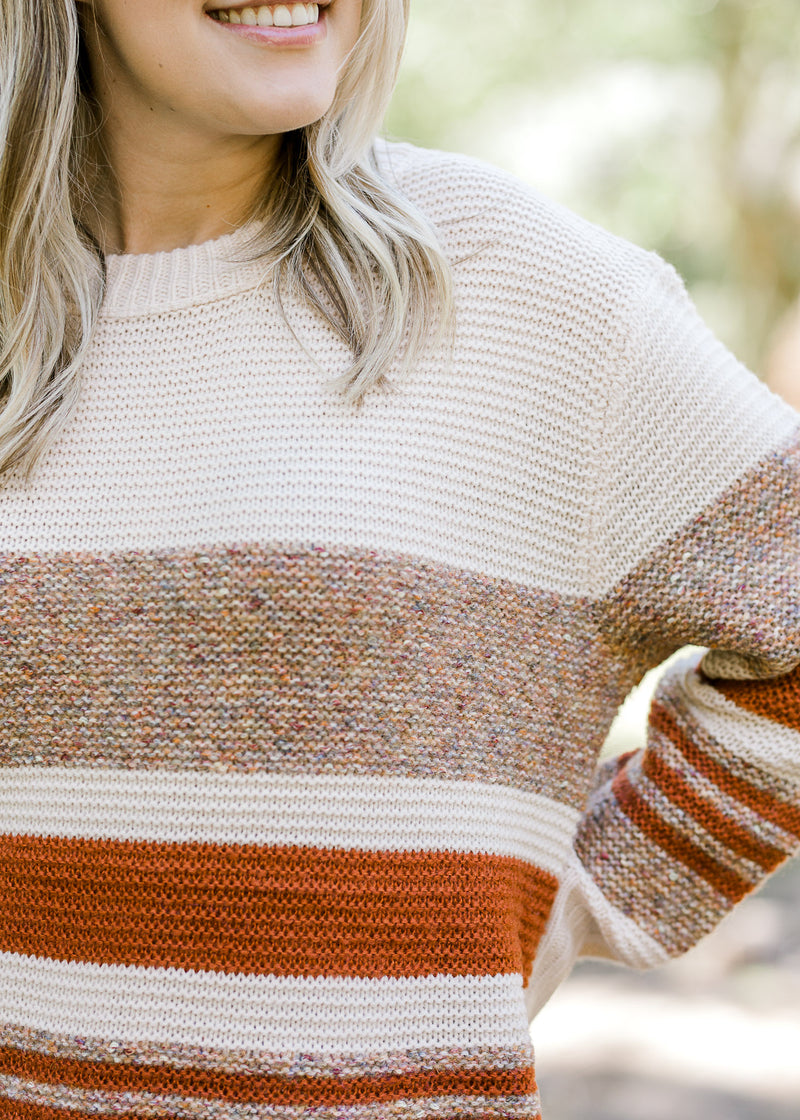 Close up of Blonde model wearing cream sweater with brown and rust stripes.