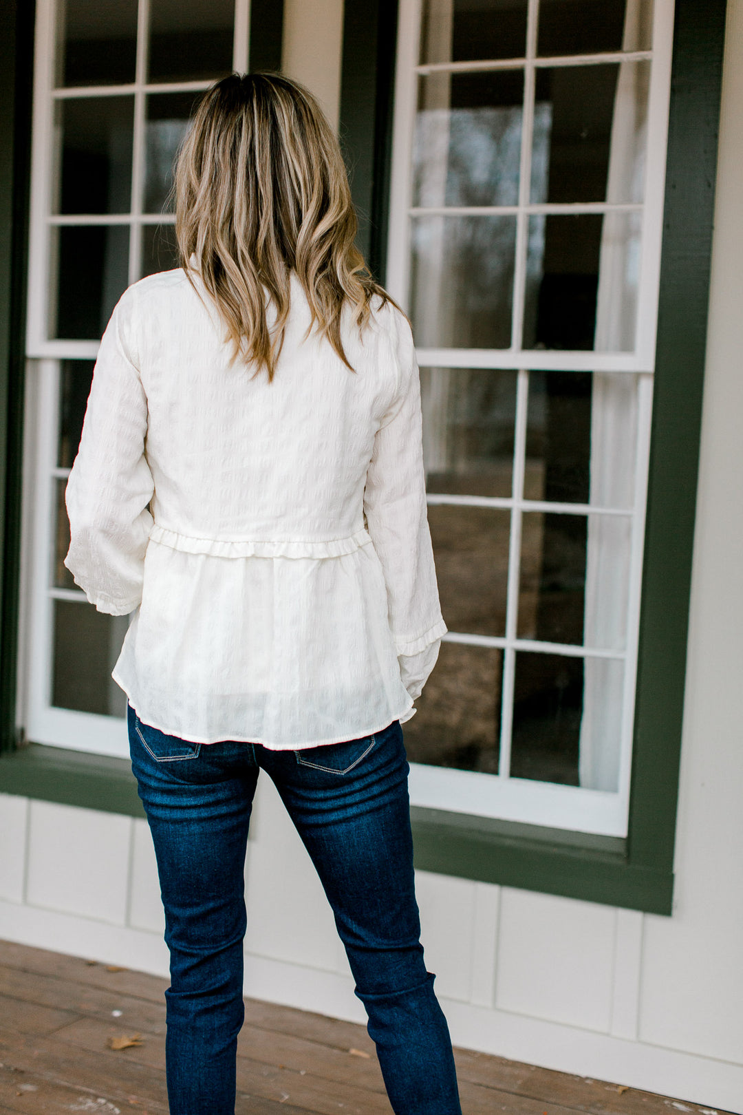 Back view of Blonde model wearing a cream top with ruffles details.