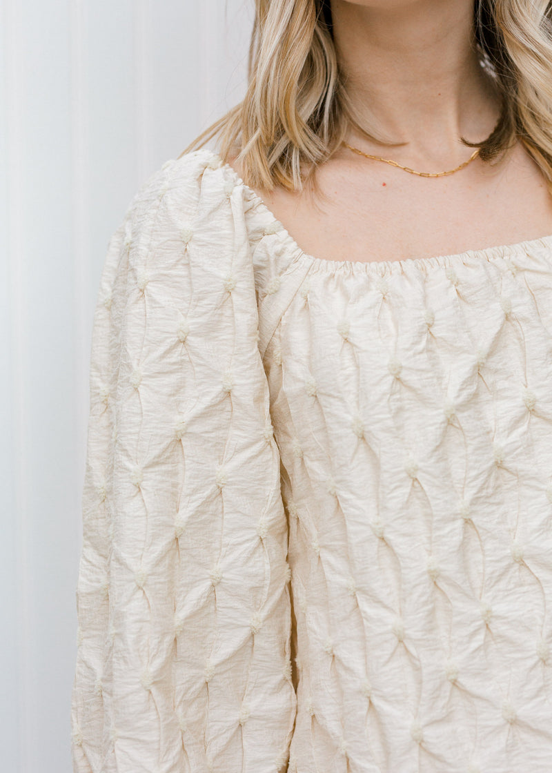 Close up view of Blonde model wearing a textured cream blouse.