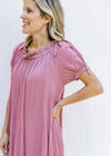 Close up view of a model wearing a rose colored dress with bubble short sleeves and a tie neck. 