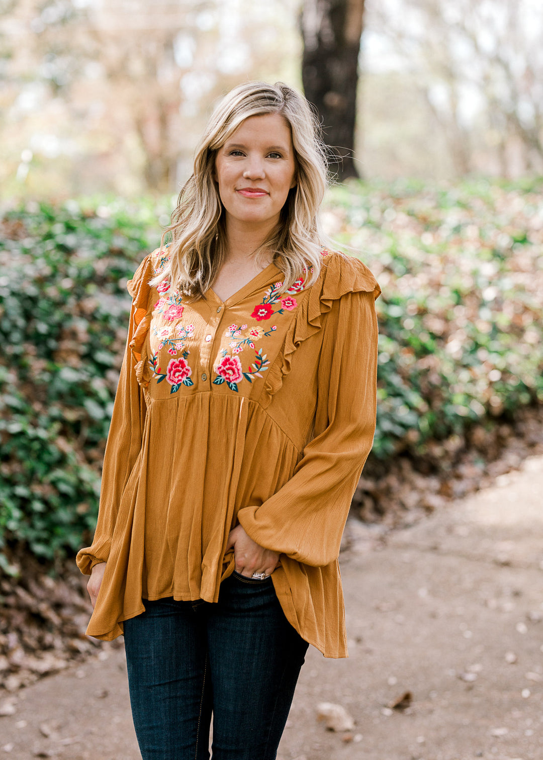 Blonde model wearing mustard, long sleeve blouse with floral embroidery.