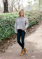 Blonde model wearing grey sweater with pink flowers, jeans and booties.