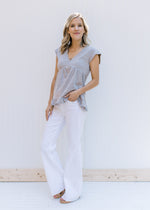 Model wearing a v-neck gray babydoll top with capped sleeves and white wide leg pants. 