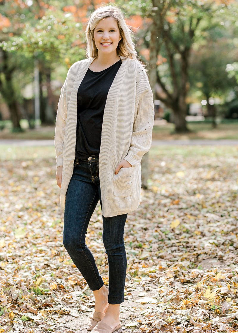Blonde model wearing cream cardigan with black top, jeans and slip on shoes.