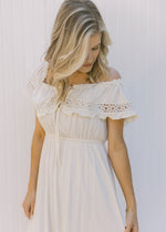 Close up view of model wearing an off the shoulder cream dress with lace detail and a tie at neck. 