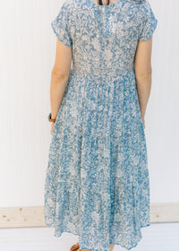 Back view of Model wearing a blue button up midi dress with an ivory pattern and a tiered design. 