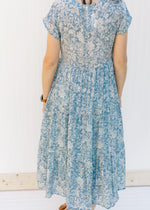 Back view of Model wearing a blue button up midi dress with an ivory pattern and a tiered design. 