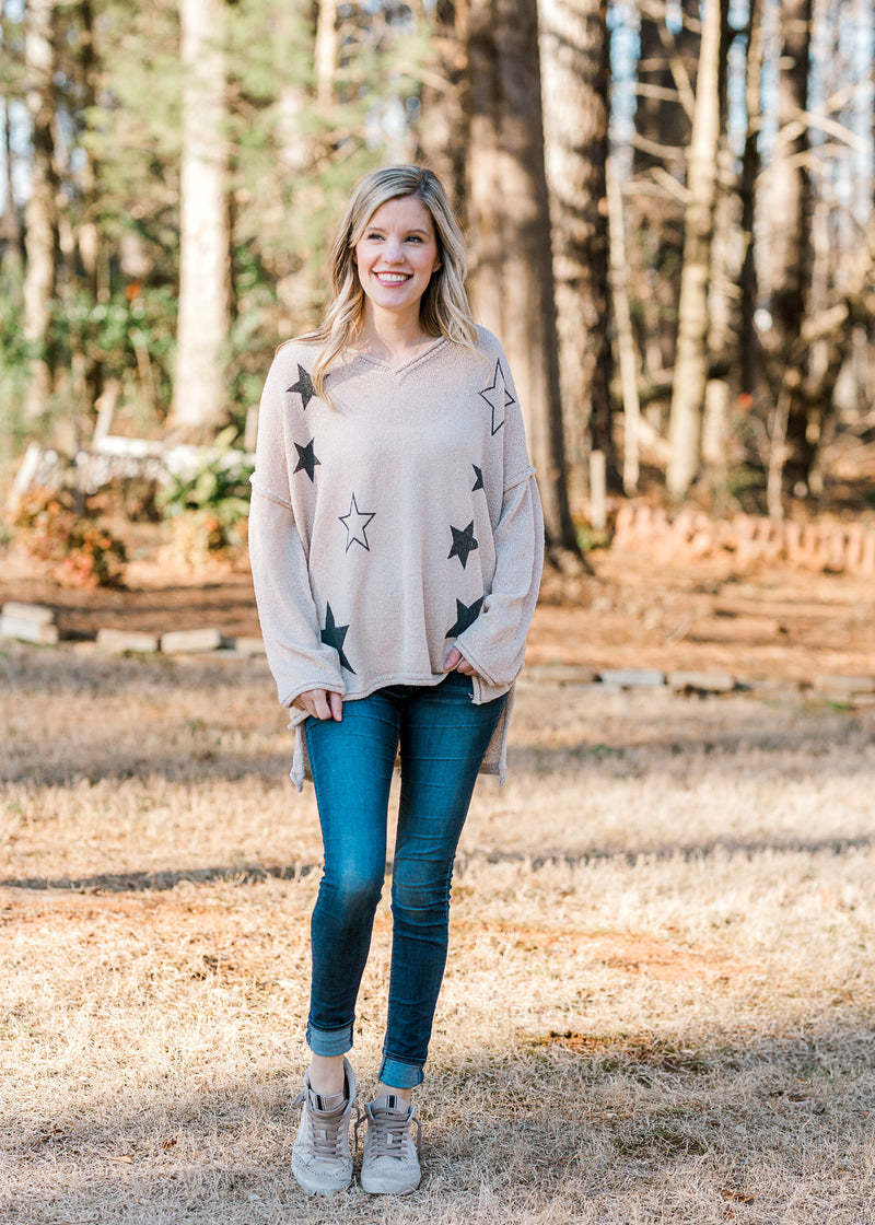 Blonde model wearing a taupe sweater with black stars, jeans, and tennis shoes.