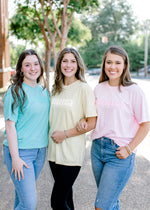 Brunette models wearing teal, yellow, and pink puff print Mama comfort color tees.