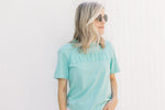 Model wearing sunglasses with a teal short sleeve tee shirt with a raised mama graphic. 