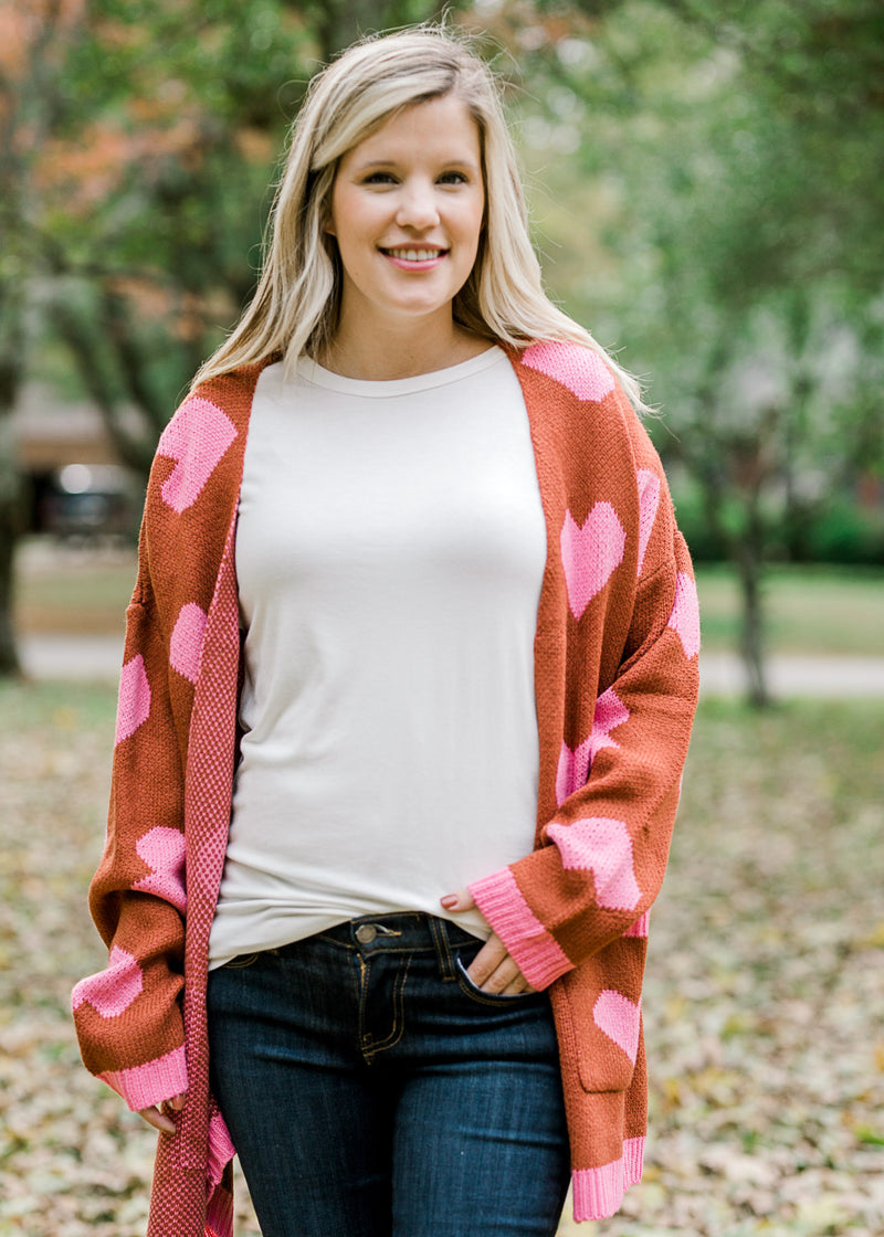 Blonde model wearing a white top, jeans and a rust cardigan with pink hearts.