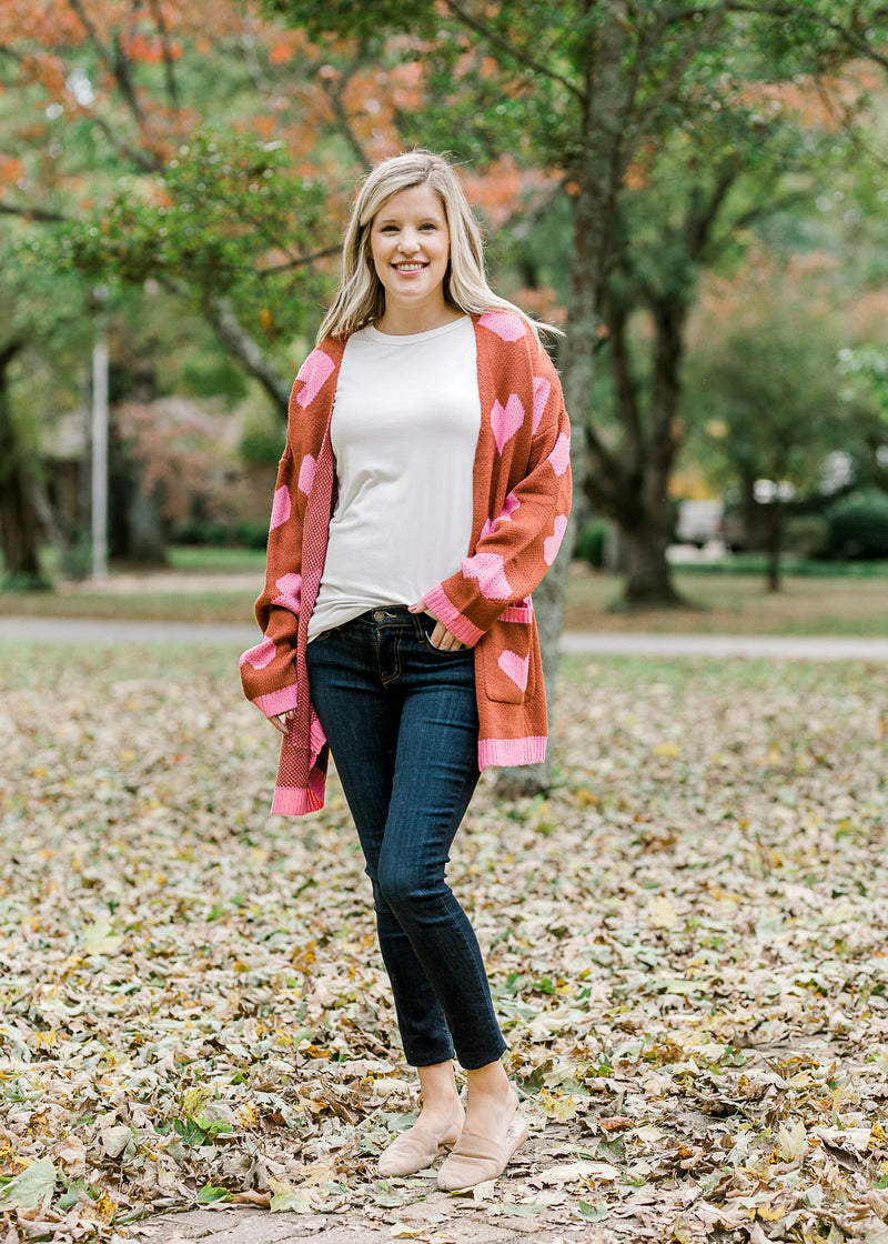 Blonde model wearing jeans, a white top with a rust cardigan with pink hearts and slip on shoes.
