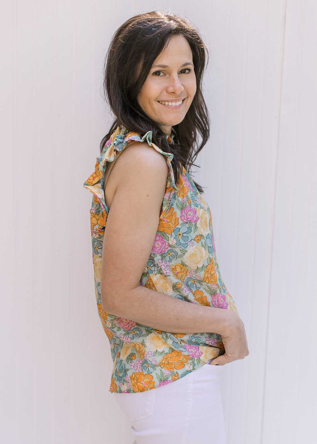 Side view of model wearing a bright floral top with a ruffle on neck and arm and open stitching . 