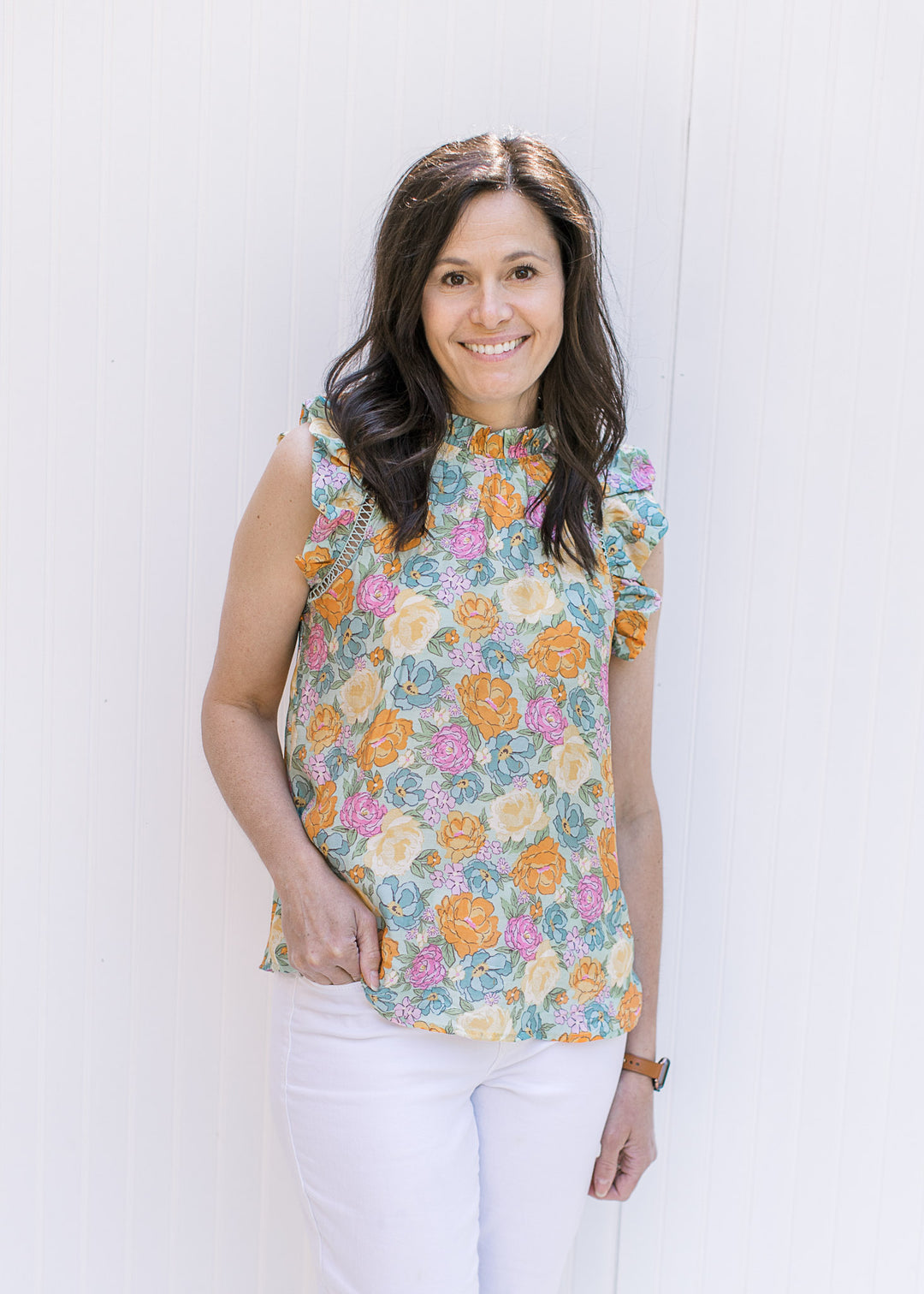 Model wearing a bright floral top with a ruffle at the neck and arm and white jeans. 