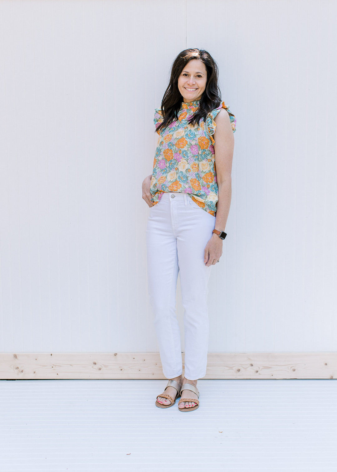 Model wearing a bright floral top with a ruffle at the neck and arm with white jeans and sandals. 