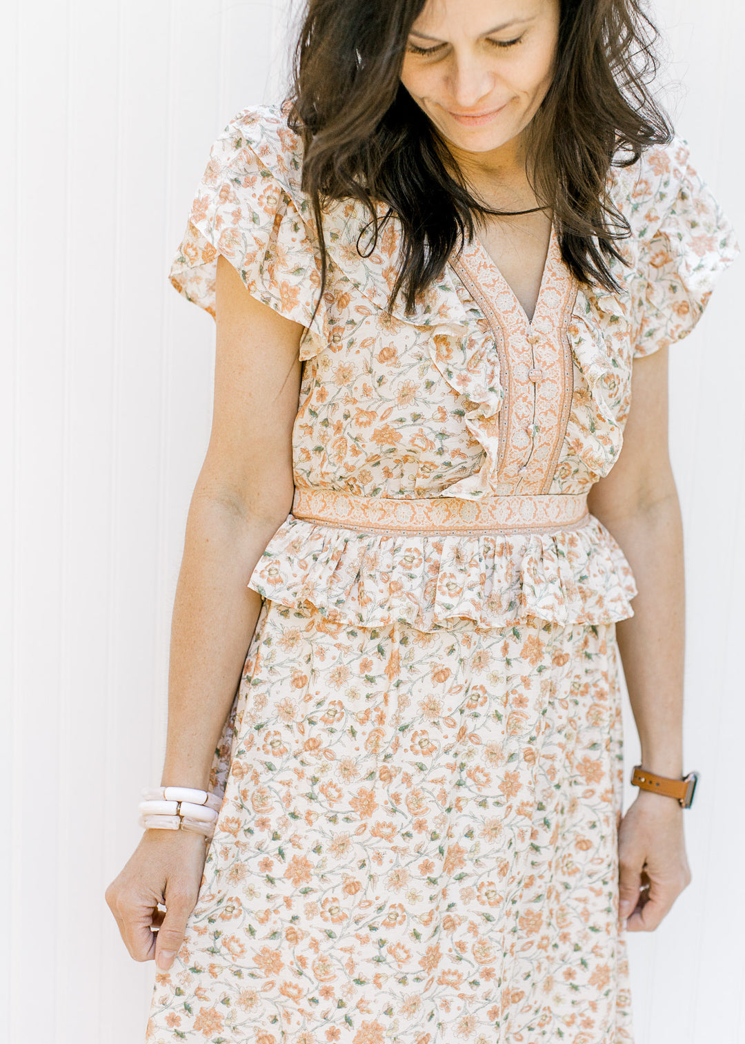 Model wearing a cream v-neck dress with a peach floral design with a ruffle and elastic at waist. 