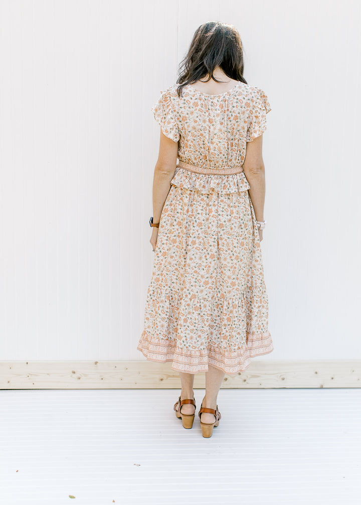 Back view of Model wearing a cream v-neck dress with a peach floral design with a ruffle at waist. 