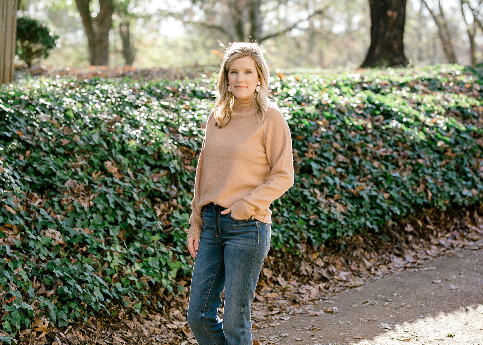 Blonde model wearing a camel color turtleneck sweater with jeans.