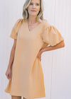 Model wearing a tan, above the knee dress with pockets and bubble short sleeves. 