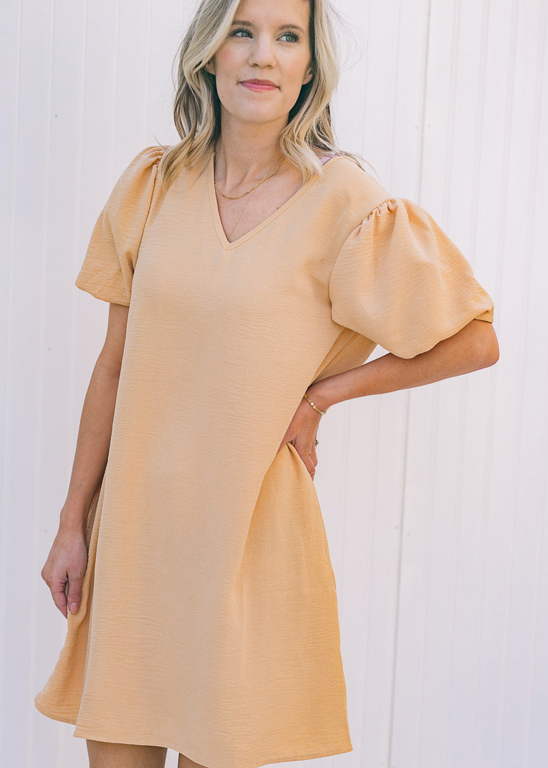 Model wearing a tan, above the knee dress with pockets and bubble short sleeves. 