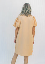 Back view of Model wearing a tan, above the knee dress with pockets and bubble short sleeves. 