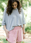 Brunette model wearing silver workout shirt and wide elastic waist shorts.