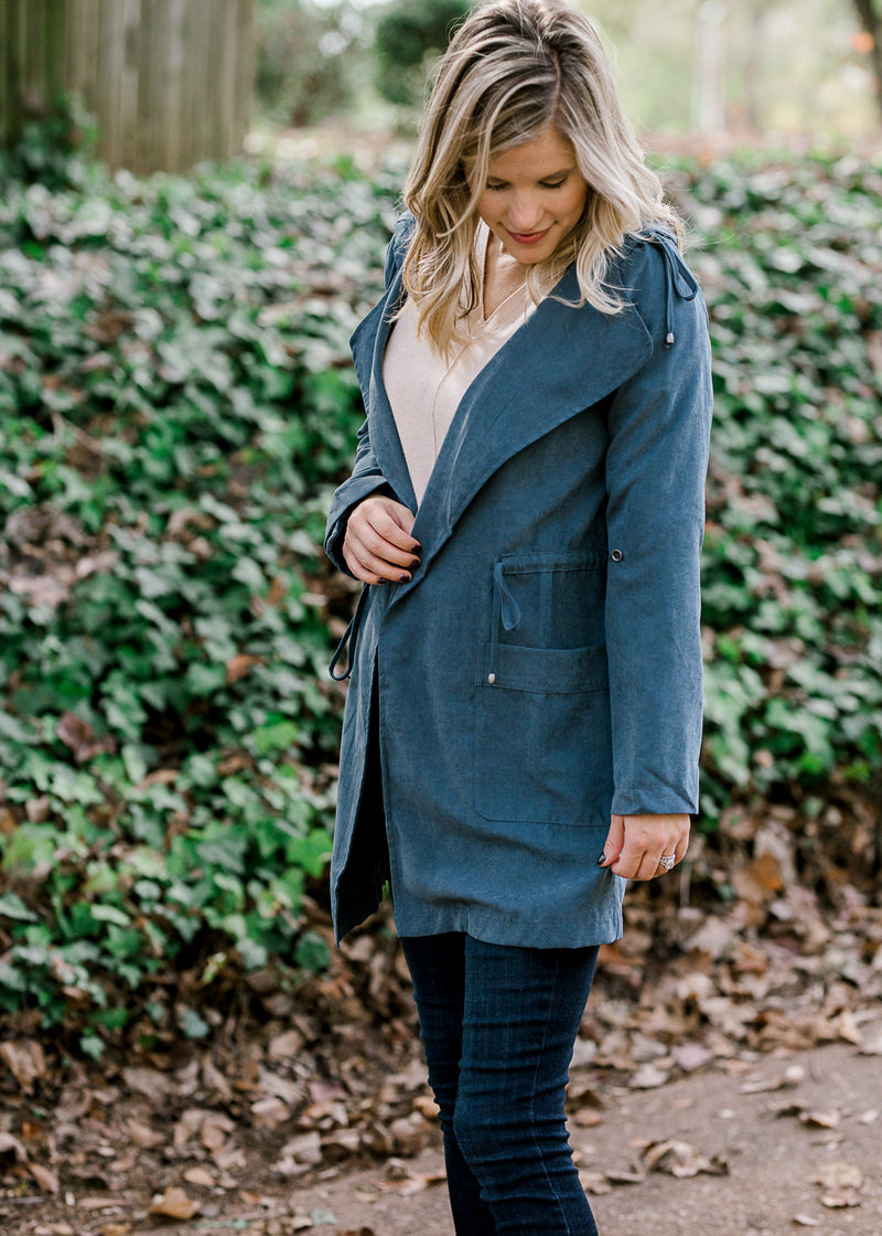 Blonde model wearing a slate blue jacket with pockets and a tie at the waist. 