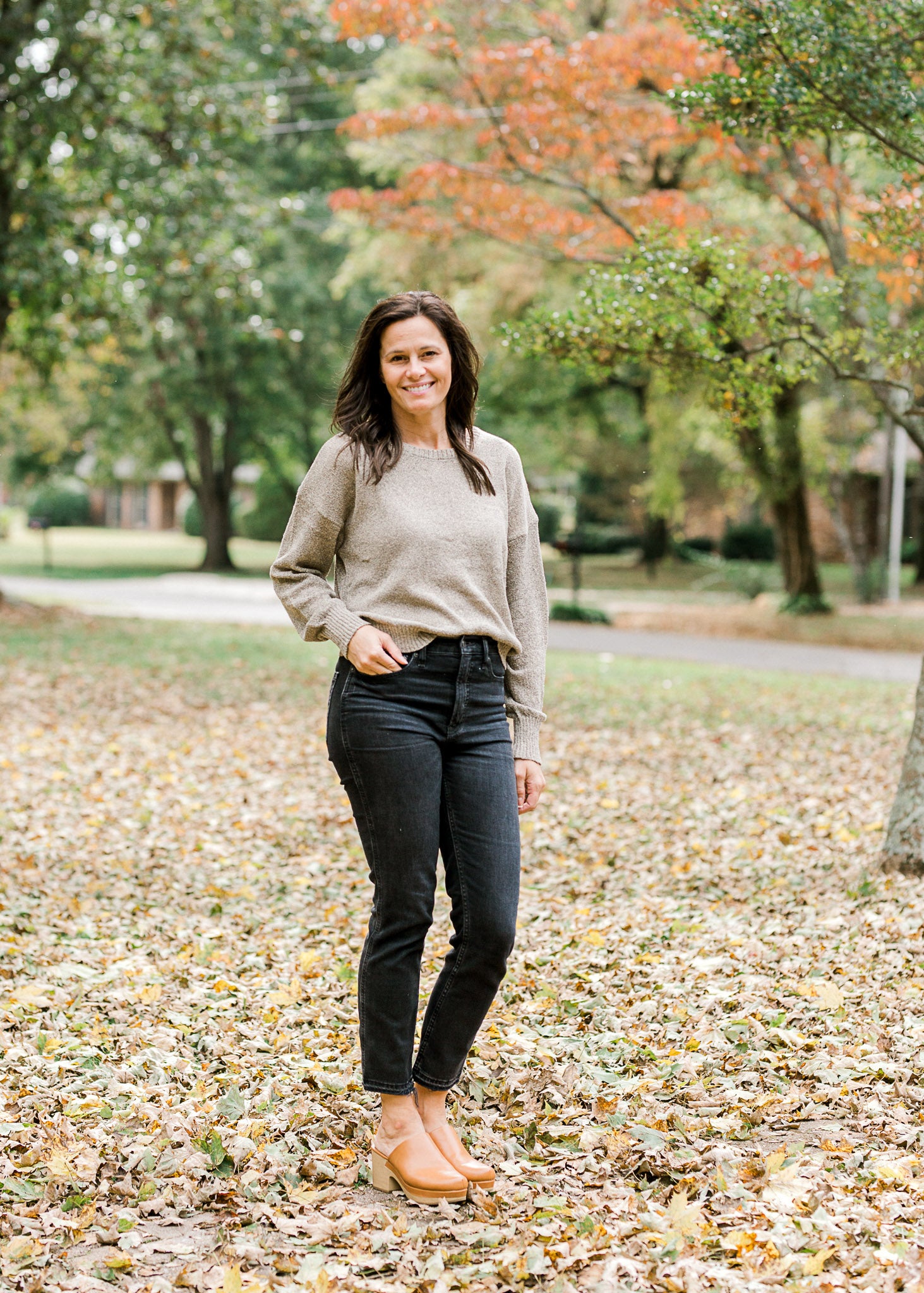 Brunette model wearing taupe sweater with gold shimmer detail with black jeans and mules.