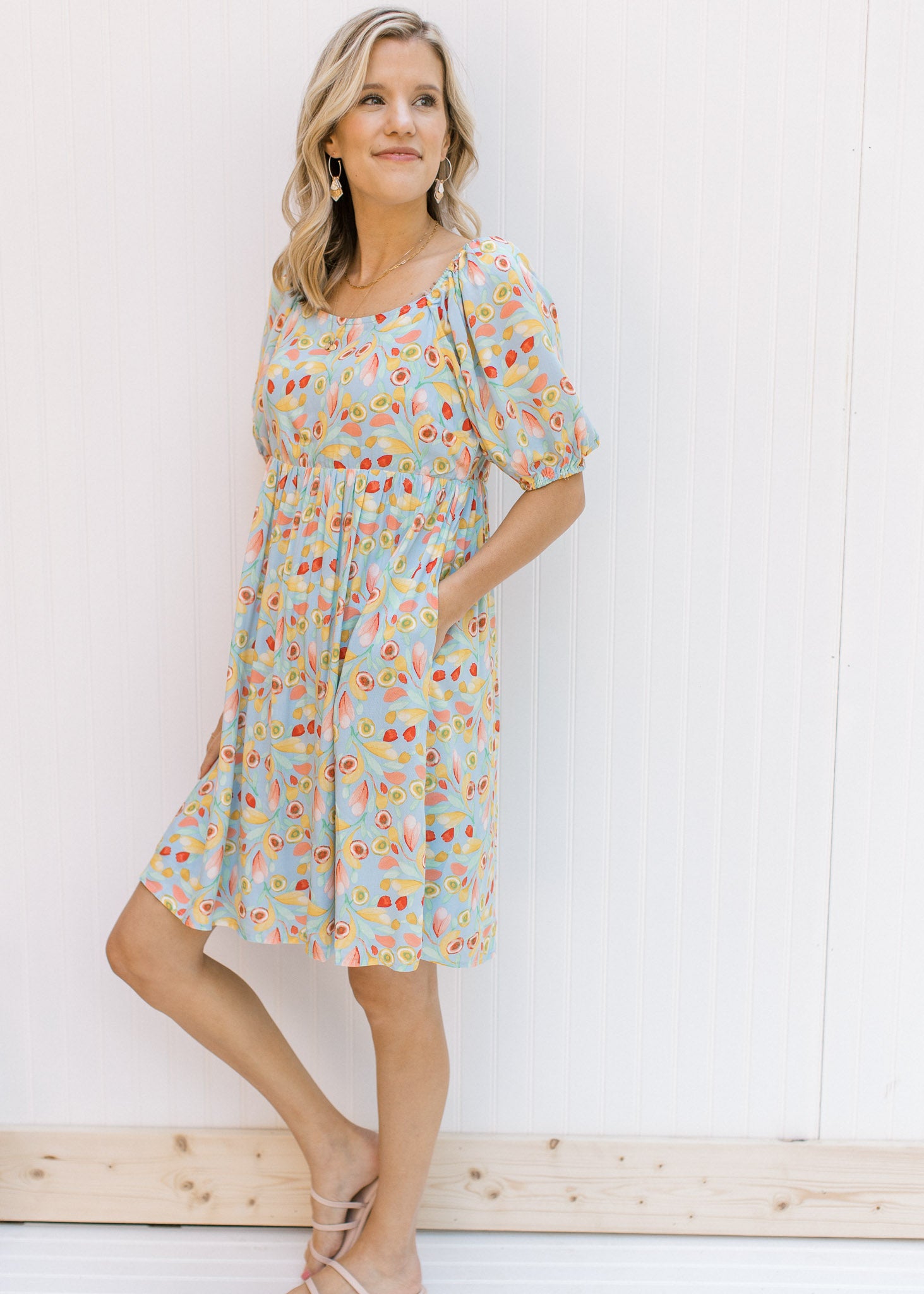 Model wearing sandals with an above the knee light blue babydoll dress with a bright floral design. 
