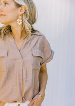 Close up view of Model wearing a taupe v-neck top with chest pockets and cuffed short sleeves. 